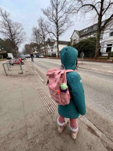 Ein Kind mit pinkem Rucksack steht im Wintermantel am Rand eines Gehwegs und schaut auf eine befahrene Straße ohne Zebrastreifen; Autos und ein Radfahrer fahren vorbei, im Hintergrund Wohnhäuser – der unsichere Schulweg wird deutlich.