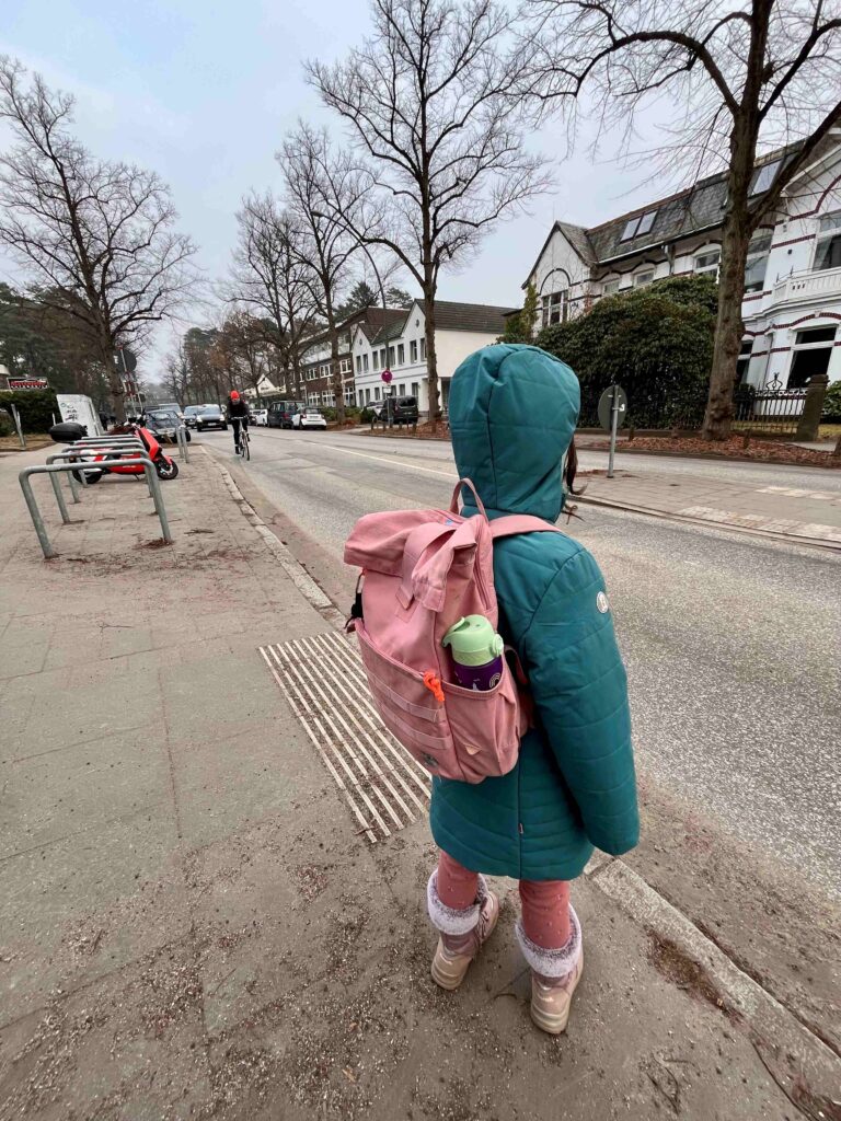 Ein Kind mit pinkem Rucksack steht im Wintermantel am Rand eines Gehwegs und schaut auf eine befahrene Straße ohne Zebrastreifen; Autos und ein Radfahrer fahren vorbei, im Hintergrund Wohnhäuser – der unsichere Schulweg wird deutlich.