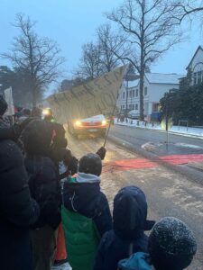 Rückansicht einer Menschenmenge aus Kindern und Erwachsenen, die bei Schneefall an einer Straße demonstrieren. Ein roter Teppich liegt quer über der Fahrbahn, während die Gruppe auf ein herannahendes Fahrzeug mit eingeschalteten Scheinwerfern blickt und ein Protestplakat hochhält.