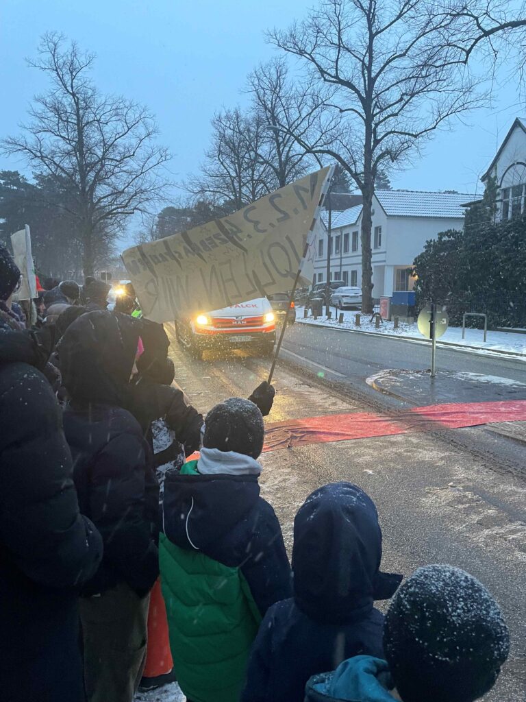 Rückansicht einer Menschenmenge aus Kindern und Erwachsenen, die bei Schneefall an einer Straße demonstrieren. Ein roter Teppich liegt quer über der Fahrbahn, während die Gruppe auf ein herannahendes Fahrzeug mit eingeschalteten Scheinwerfern blickt und ein Protestplakat hochhält.